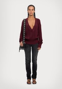 Suéter de punto burdeos con un profundo escote en V, con textura en las mangas, combinado con jeans ajustados negros y sandalias negras con tiras, llevando un bolso pequeño.