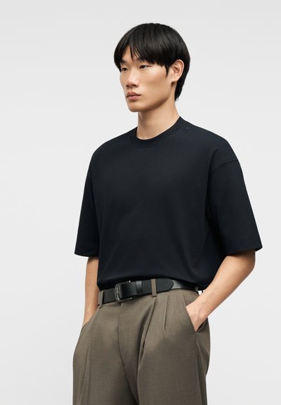 Νεαρός άνδρας με μαύρο υπερμεγέθες T-shirt που είναι μπουσουλιασμένο σε καφέ ψηλόμεσα παντελόνια με πιέτες και μαύρη ζώνη, τα χέρια στις τσέπες.