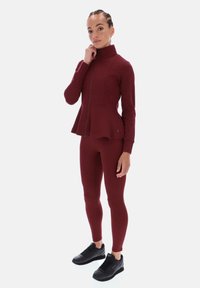 Žena stojí a má na sobě přiléhavou vínovou bundu na zip s peplum lemem, ladící legíny a černé tenisky, na jednobarevném pozadí.