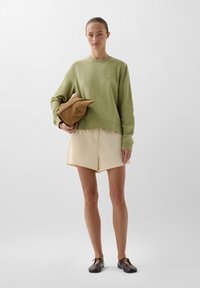 Lys grøn sweatshirt med ribbede manchetter og kant, beige shorts og en brun taske. Modellen har mørke sko på og står foran en ensfarvet baggrund.