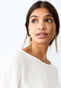 Bílý top s jemnými proužky ve textuře, zaobleným výstřihem a volným střihem. Zlaté náušnice s třásněmi přidávají kontrast a detail.