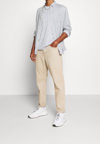 Pantaloni beige, t-shirt grigio chiaro e strutturato e camicia celeste con bottoni. Scarpe da ginnastica bianche con dettagli neri completano l'outfit.
