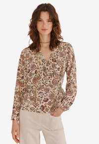 Langærmet bluse i beige stof med blomstertryk i rød, gul og grøn. Har V-hals og taljebånd for justerbar pasform.