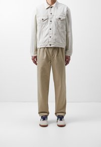 Mand iført en off-white denimjakke, beige bukser og hvide sneakers med brune såler, stående foran en ensfarvet hvid baggrund.