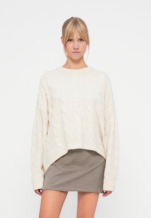 Pull crème en tricot torsadé avec des manches longues et un ourlet asymétrique. Associé à une mini-jupe à carreaux, créant un look superposé.