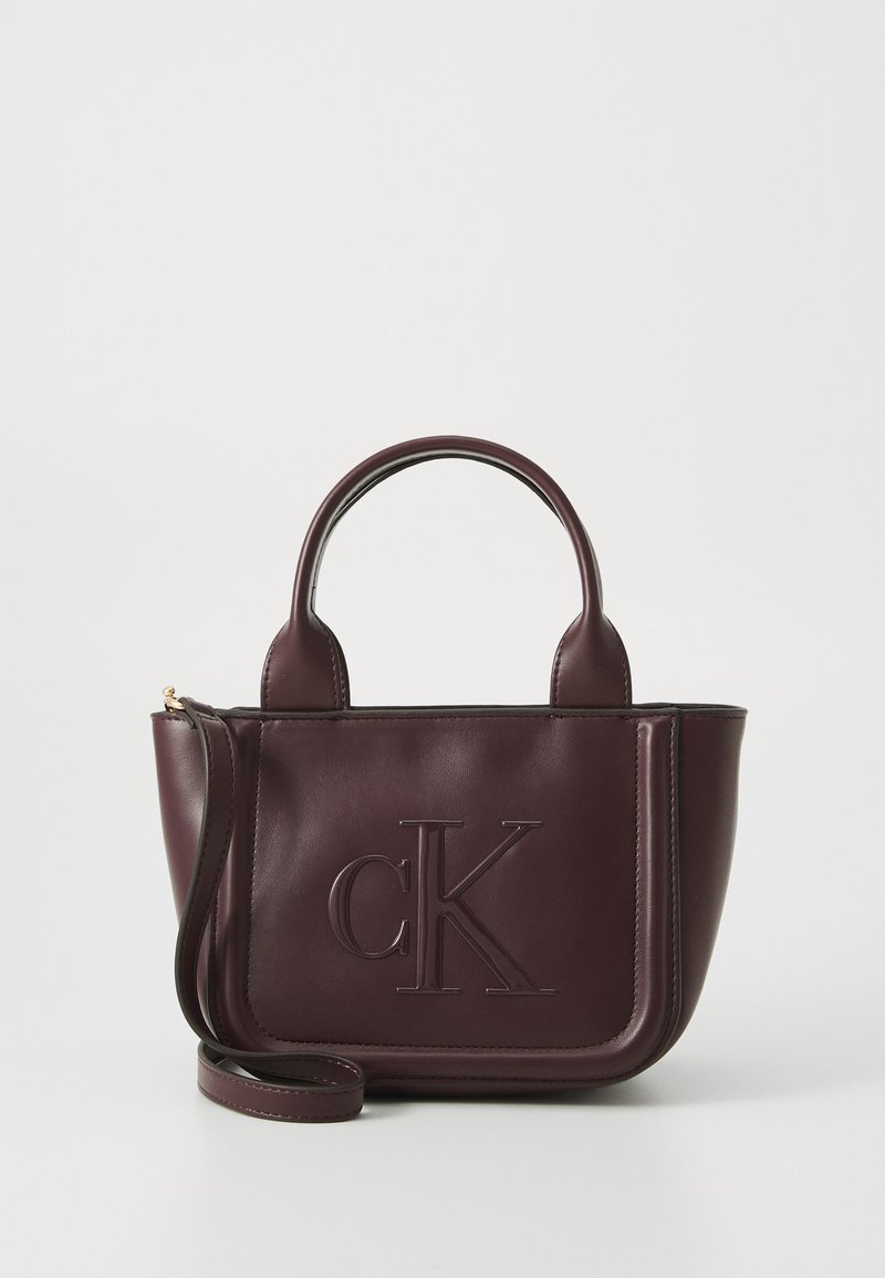 Borsa in pelle bordeaux con logo "cK" in rilievo, doppi manici corti, tracolla staccabile e forma strutturata. Texture liscia.
