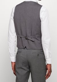 Szary wełniany kamizel z dopasowanym krojem, z paskiem regulacyjnym z tyłu i teksturowaną powierzchnią. W zestawie z szarymi spodniami i białą koszulą.
