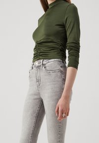 Top de manga larga verde oscuro con detalles fruncidos en la cintura, combinado con jeans de mezclilla gris claro que cuentan con un cierre de botón y bolsillos delanteros.
