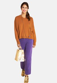 Frau lächelt, trägt einen braunen Pullover, lila Hosen, goldene Sneaker, hält eine goldene Handtasche, ist mit einer langen Kette und Ohrringen geschmückt und geht vorwärts.