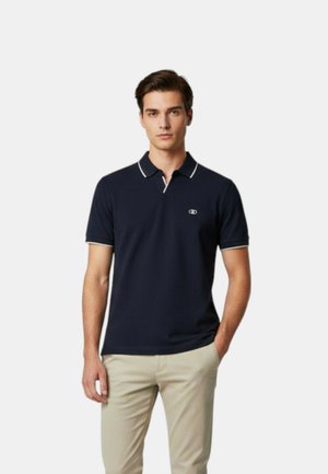 Giovane uomo in piedi con una mano in tasca, indossa una polo blu navy con bordi bianchi e pantaloni beige su sfondo semplice.