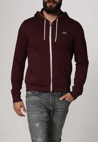 Burgundyröd zip-up hoodie med en framficka, vita dragsnören och en liten logodetalj. Tillverkad av mjukt material med en slät textur.