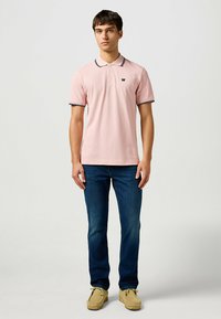 Camisa de polo rosa con cuello, tres botones y ribete azul marino en las mangas. Llevada con jeans azules y zapatos amarillos. Logo simple en el pecho.