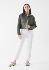 Giacca cropped in pelle verde con due tasche frontali, abbinata a jeans bianchi e zoccoli beige; texture liscia, dettagli con bottoni.
