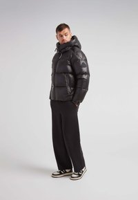 Giacca a vento nera con colletto alto e cappuccio, caratterizzata da una texture lucida, baffle orizzontali e una toppa con logo sulla manica. Abbinata a pantaloni neri.