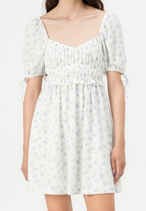 Kobieta ubrana w białą mini sukienkę w jasnoniebieski kwiatowy wzór, z bufiastymi rękawami, kwadratowym dekoltem i marszczoną talią z falbaną.