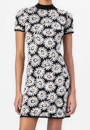 Robe courte noire avec motifs de marguerites blanches et rose clair, manches courtes et col haut portée par une personne debout.