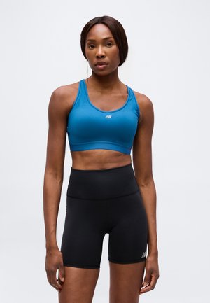 Femme en forme portant un soutien-gorge de sport bleu et un short de sport taille haute noir, debout devant un fond clair uni.