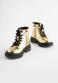 Goldfarbene metallische Ankle Boots mit schwarzen Schnürsenkeln und einer dicken schwarzen Sohle. Sie verfügen über eine glatte Oberfläche und einen gepolsterten Kragen für zusätzlichen Komfort.
