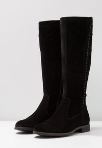 Botas de ante negras hasta la rodilla con un acabado suave, detalle de costura lateral y un tacón bajo y rectangular. Diseño de punta cuadrada.