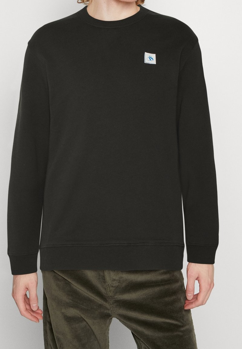 Sudadera negra de cuello redondo fabricada en algodón, con mangas largas y un pequeño parche de logo en azul y blanco en el pecho. Acabado de tela texturizada.