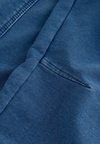 Artimų planas, kuriame matoma sulankstyta mėlyna džinsinė medžiaga, išryškinanti detalų tekstūrą ir siūles kišenės srityje.