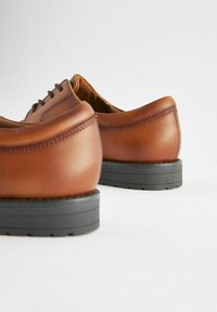 Chaussures en cuir marron avec une texture lisse, dotées de semelles en caoutchouc gris et de lacets traditionnels. Le design comprend des détails de couture subtils.