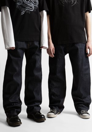 Dwóch mężczyzn w luźnych ciemnych dżinsach i czarnych koszulkach z nadrukiem, jeden z białymi, długimi rękawami pod spodem, stojących obok siebie.