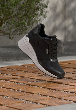 Baskets en cuir noir avec semelle épaisse blanche et logo "DKNY", présentées en extérieur sur un sol carrelé avec un fond de plantes vertes et un mur en béton.