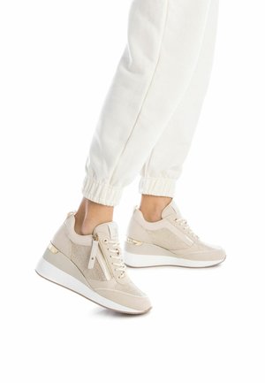 Zapatillas deportivas beige con paneles de malla, acentos en dorado y diseño de suela cuña. Cordones y detalle de cremallera lateral visibles.