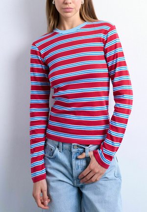 Femme portant une chemise rouge à manches longues avec des rayures horizontales bleues et blanches et un jean bleu clair, debout devant un fond blanc uni.