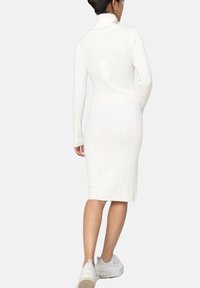 Robe en tricot côtelé blanche avec un col montant, des manches longues et une silhouette ajustée de type bodycon, atteignant le mi-mollet. Portée avec des baskets blanches.