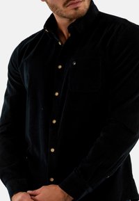 Chemise en velours côtelé vert foncé avec des manches longues, un col boutonné, des boutons dorés et une poche poitrine. Présente une texture subtile et un design classique.