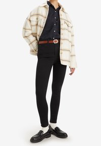 Camisa chaqueta de cuadro en crema y naranja oversized sobre una camisa negra de botones, combinada con jeans skinny negros y mocasines negros con calcetines blancos.