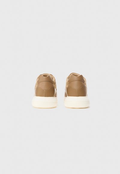 Un paio di sneakers beige con suole bianche viste da dietro su uno sfondo grigio chiaro uniforme.