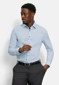 Hellblau gemustertes Hemd mit kleinen Punkten, langen Ärmeln, Button-Down-Kragen und dunklen Knöpfen, kombiniert mit dunkelgrauen Hose und einem schwarzen Gürtel.