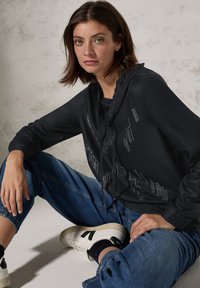 Haut à capuche noir avec un design texturé, présentant des accents argentés, associé à un jean bleu déchiré et des baskets blanches avec des détails noirs.