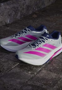 Paire de chaussures de course Adidas Adizero blanches et roses avec des lacets blancs sur une surface en pierre texturée.