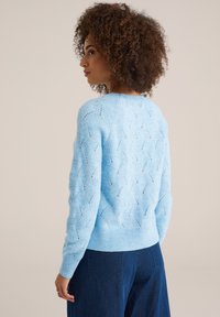Lichtblauwe gebreide trui met een golvenpatroon en opengewerkte details, met geribde manchetten en zoom. Gelangd met blauwe denim broek.