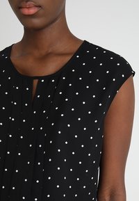 Gros plan sur une personne portant un haut sans manches noir avec des pois blancs et un petit détail de décolleté en forme de clé.