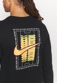 Schwarzes Langarm-T-Shirt mit einem großen orangen Nike-Logo, ergänzt durch gelbe grafische Elemente und Text zu Fußball, vor einem schlichten Design.