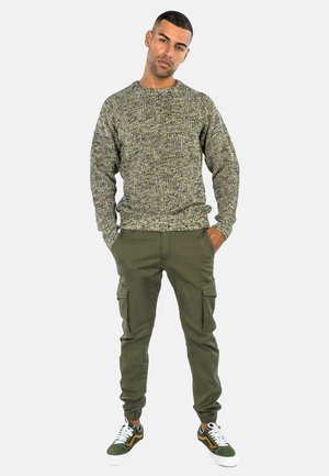 Maglione grigio textured, pantaloni cargo verdi con tasche e sneakers sportive verdi e bianche. Outfit casual mostrato su un modello maschile.