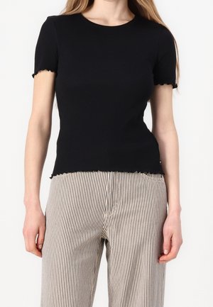 Kobieta ubrana w dopasowaną czarną bluzkę z krótkim rękawem z falbankami oraz wysokie spodnie w pionowe czarno-białe paski.