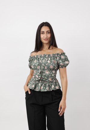 Femme aux longs cheveux foncés portant un chemisier vert fleuri à épaules dénudées et un pantalon noir, debout avec une main dans la poche sur un fond uni.