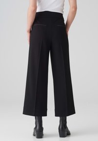 Pantalons larges noirs avec une texture lisse, présentant une taille haute, des poches arrières et des lignes épurées. Associés à des bottes à talons noires.