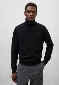 Schwarzer Rollkragenpullover aus weichem Strickmaterial, mit geripptem Kragen und Bündchen, kombiniert mit grauen Hosen mit seitlichen Taschen.