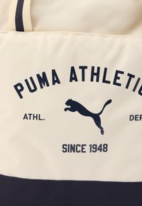 Μπεζ τσάντα υφασμάτινη tote με μπλε-γκρι βάση, με την επιγραφή «PUMA ATHLETICS» και λογότυπο πούμα σε πήδημα σε μπλε-γκρι χρώμα. Επιφάνεια με υφή και δύο χειρολαβές.