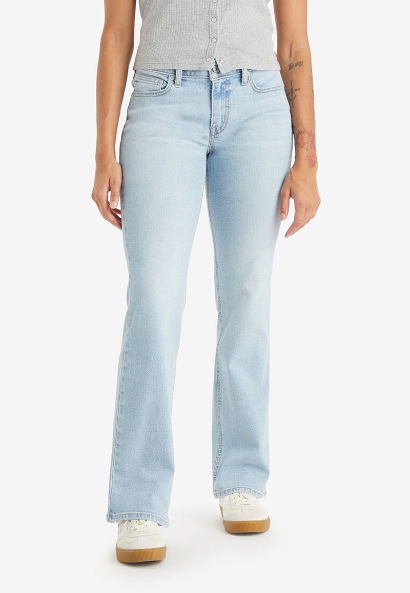 Femme portant un jean bootcut bleu clair, des baskets blanches et un haut sans manches gris à boutons, se tenant sur un fond blanc.