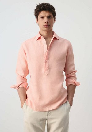 Jeune homme aux cheveux foncés portant une chemise rose clair à manches longues retroussées et un pantalon beige clair, debout les mains dans les poches.