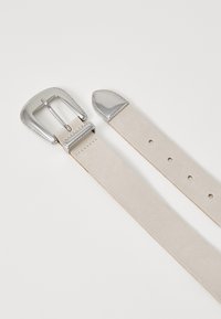 Ceinture en cuir beige clair avec une boucle rectangulaire en argent et un bord décoratif. Texture lisse avec des trous perforés et une pointe effilée pour le réglage.