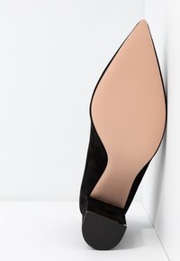 Chaussure en daim noir à bout pointu vue de la semelle, alternant une semelle lisse beige pâle avec un talon bloc noir bas sur fond blanc.